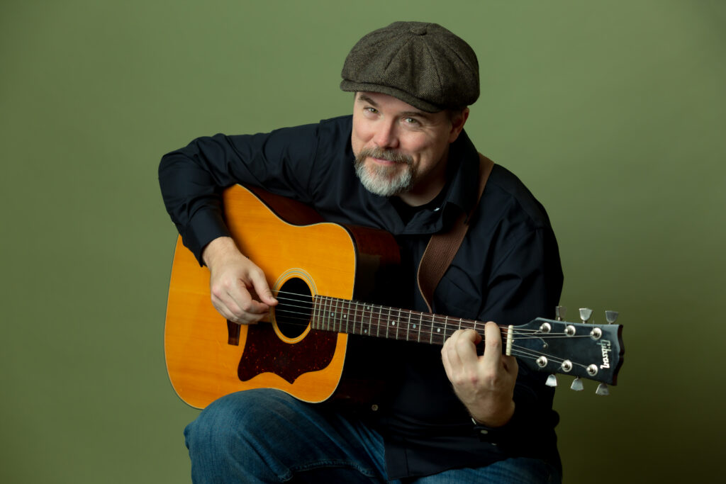 Photo of Paul Strickland playing a guitar in front of a green background